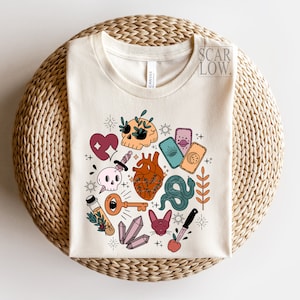 Op de afbeelding: Een wit T-shirt met een kleurrijk grafisch ontwerp met een schedel, hart, slang, sleutel, kristallen en tarotkaarten. Het ontwerp is gecentreerd op het shirt en wordt omringd door een witte rand.