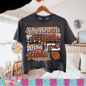 May include: Dark gray t-shirt with a basketball-themed graphic. The design includes the phrases "Hoop there it is," "It's Gameday," "Defense Offense," and "Basketball Season," along with a basketball illustration. The shirt is hanging on a wooden hanger.