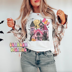 May include: A white t-shirt with a pink and black haunted house graphic. The graphic includes a full moon, bats, and the word "BOO" in colorful letters.