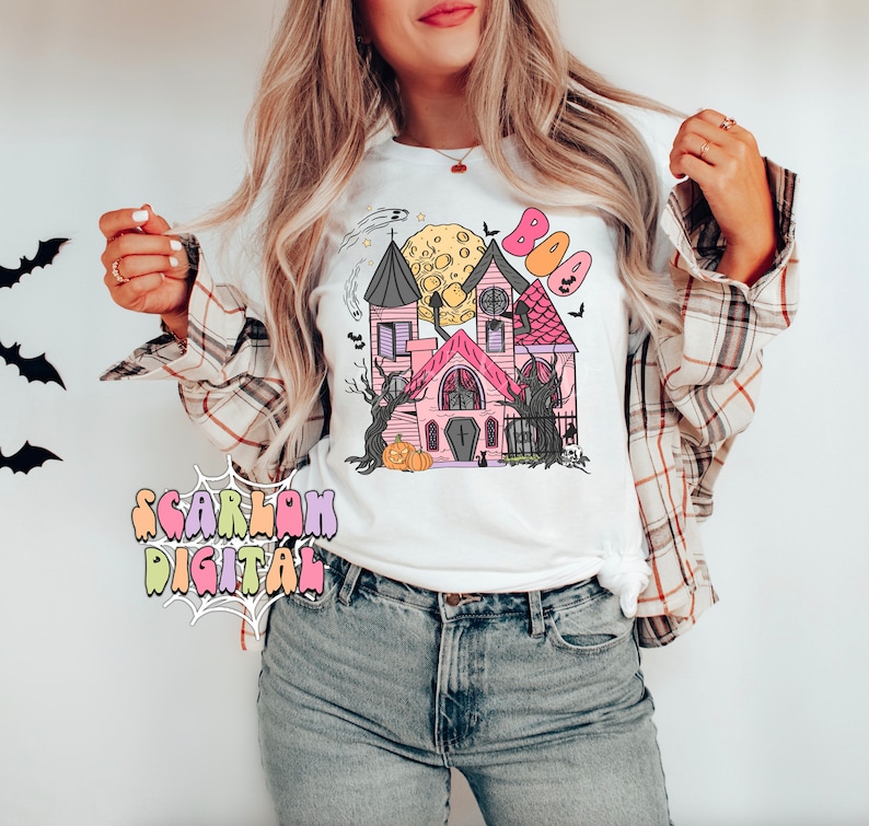 May include: A white t-shirt with a pink and black haunted house graphic. The graphic includes a full moon, bats, and the word "BOO" in colorful letters.