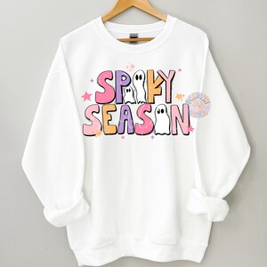 Op de afbeelding: Witte trui met een roze, paarse en oranje afbeelding met de tekst "Spooky Season" met geesten en sterren.