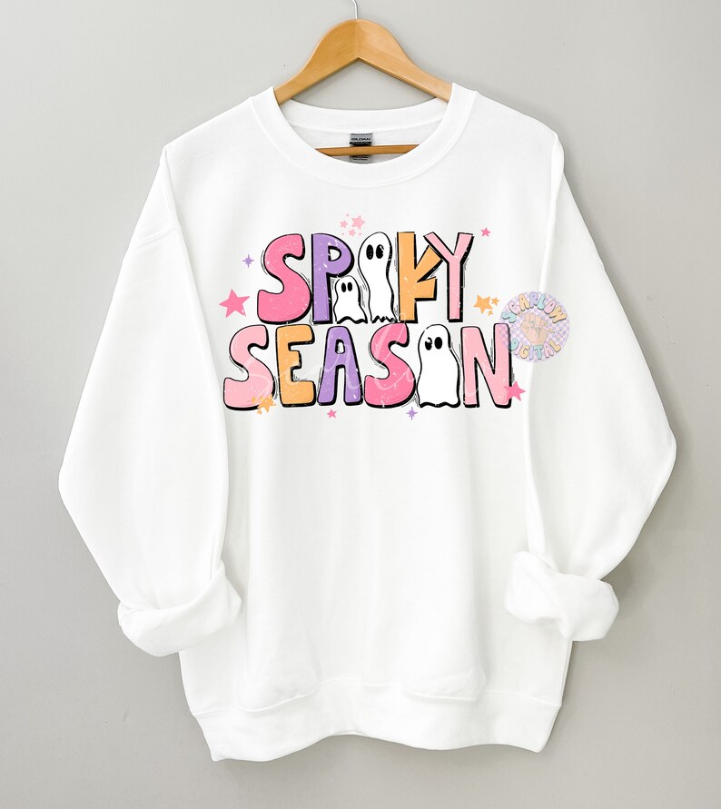 Op de afbeelding: Witte trui met een roze, paarse en oranje afbeelding met de tekst "Spooky Season" met geesten en sterren.