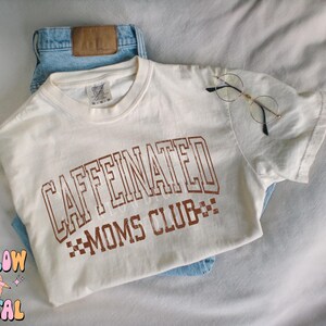 K&ouml;nnte beinhalten: Ein wei&szlig;es T-Shirt mit braunem Text, der "CAFFEINATED MOMS CLUB" lautet, mit einem karierten Muster unter dem Text.