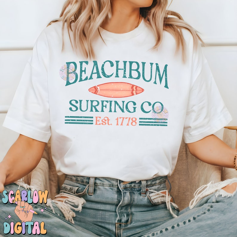 May include: White t-shirt with a teal and pink graphic design that says "Beachbum Surfing Co. Est. 1778". The graphic includes a surfboard and floral accents.