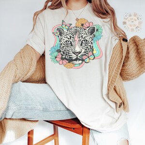 May include: A white t-shirt with a graphic of a leopard's face surrounded by flowers and butterflies. The leopard has a lightning bolt on its forehead. The flowers are in shades of pink, yellow, and orange. The butterflies are blue and pink.