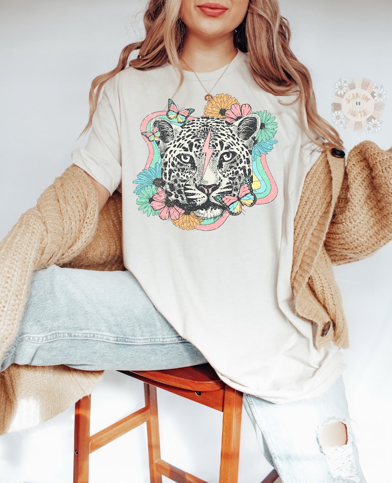 May include: A white t-shirt with a graphic of a leopard's face surrounded by flowers and butterflies. The leopard has a lightning bolt on its forehead. The flowers are in shades of pink, yellow, and orange. The butterflies are blue and pink.