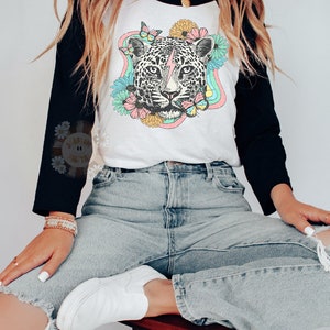 May include: A white and black baseball tee shirt with a colorful graphic of a leopard with a lightning bolt on its forehead. The leopard is surrounded by flowers and butterflies. The shirt is being worn by a person with long brown hair.