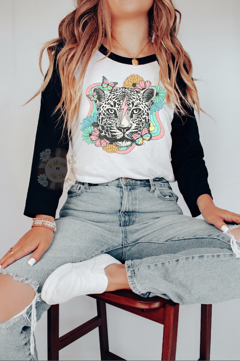 May include: A white and black baseball tee shirt with a colorful graphic of a leopard with a lightning bolt on its forehead. The leopard is surrounded by flowers and butterflies. The shirt is being worn by a person with long brown hair.