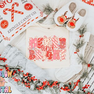 Op de afbeelding: Een cr&egrave;mekleurig T-shirt met de woorden "Merry and Bright" in roze en rood, omringd door feestelijke decoraties. De afbeelding bevat een kussen met zuurstokken, rendierpantoffels en een slinger van rode bessen, wat een vakantiethema cre&euml;ert.