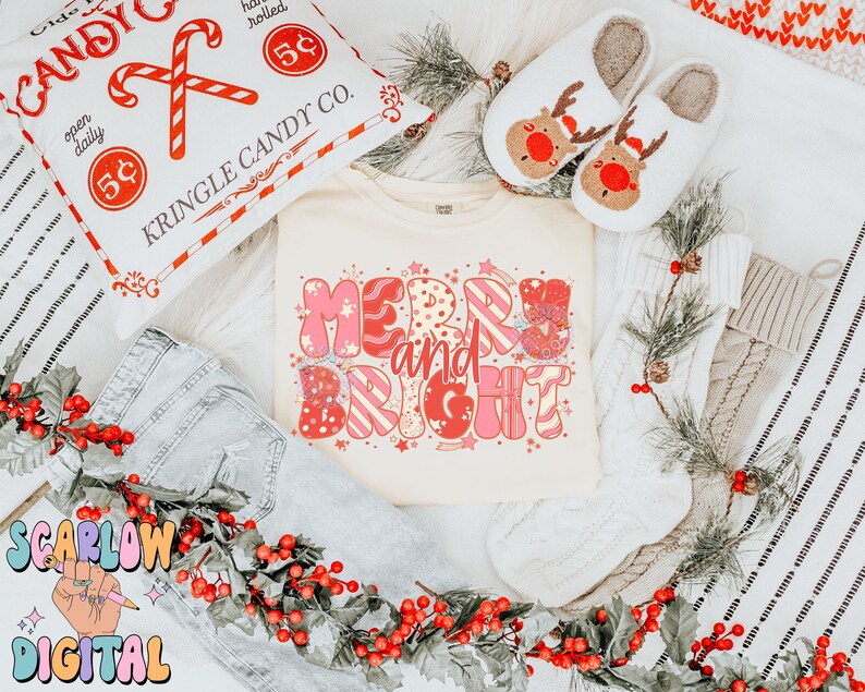 Op de afbeelding: Een cr&egrave;mekleurig T-shirt met de woorden "Merry and Bright" in roze en rood, omringd door feestelijke decoraties. De afbeelding bevat een kussen met zuurstokken, rendierpantoffels en een slinger van rode bessen, wat een vakantiethema cre&euml;ert.
