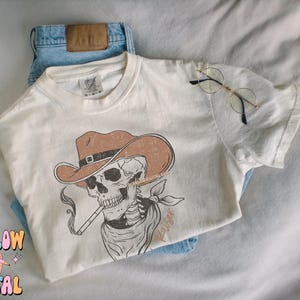 May include: A white t-shirt with a graphic of a skeleton wearing a cowboy hat and smoking a pipe. The text "Rouser" is printed on the shirt.