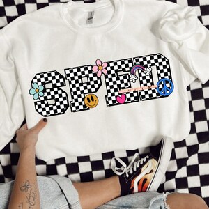 May include: White sweatshirt with the word "SPED" in black and white chequered font. The letters are decorated with colourful flowers, a rainbow, a peace sign, a smiley face, and a heart.