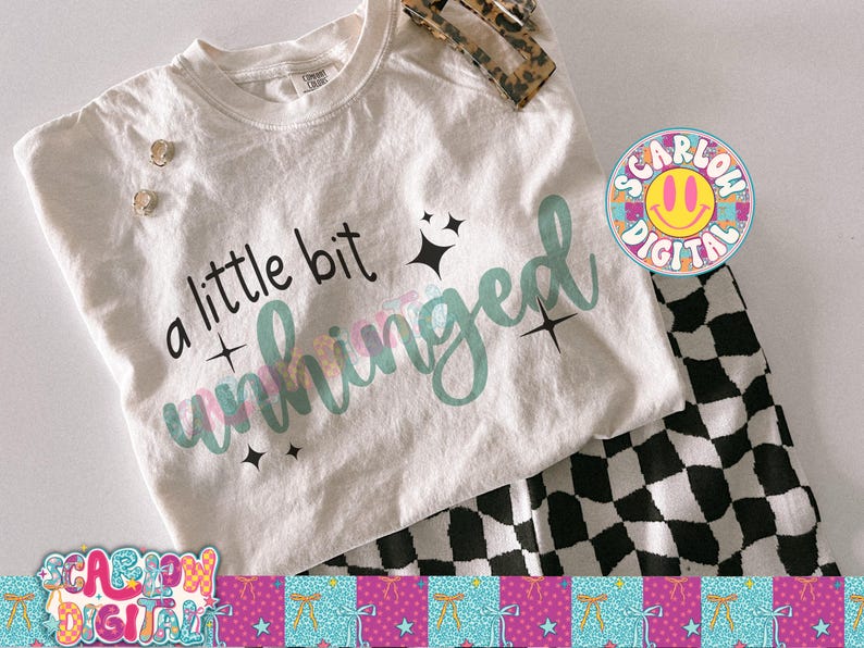 K&ouml;nnte beinhalten: Ein cremefarbenes T-Shirt mit dem Schriftzug "a little bit unhinged" in schwarzer und teal-farbener Schrift, akzentuiert mit Sternen. Das Shirt wird mit einer schwarz-wei&szlig; karierten Hose und einem Haaraccessoire mit Leopardenmuster kombiniert.