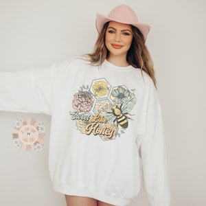 May include: White sweatshirt with a graphic of a bee and flowers in a honeycomb design. The text "Sweet Like Honey" is printed below the graphic.