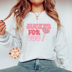 Może przedstawiać: Szara bluza z napisem "SUCKER FOR YOU" w r&oacute;żowych i białych literach. Obok napisu znajduje się r&oacute;żowy lizak w kształcie serca.