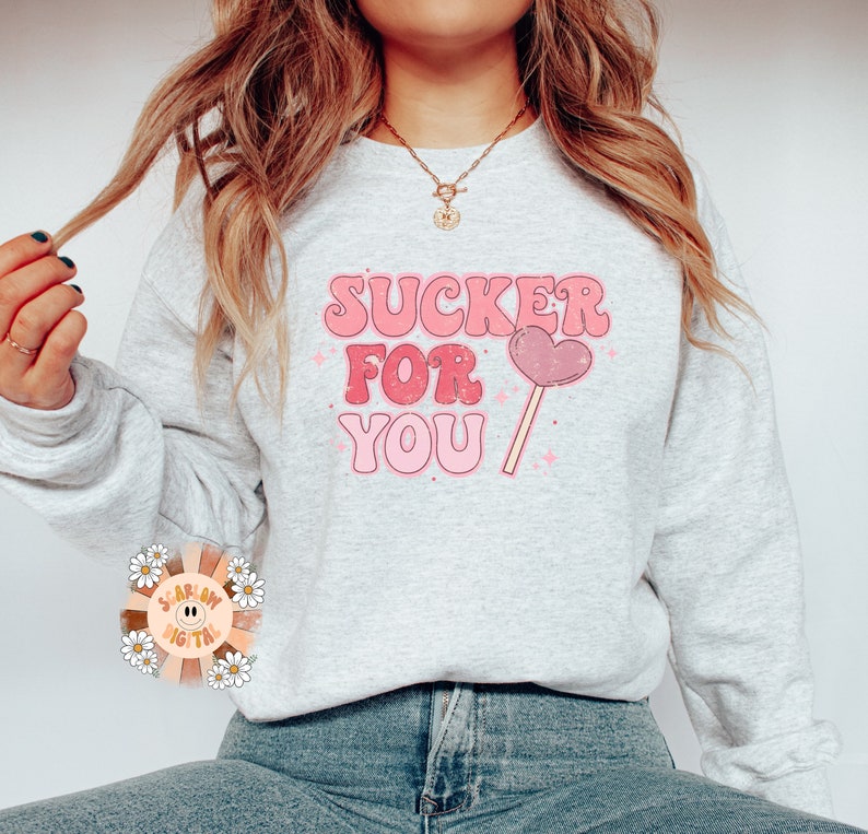 Może przedstawiać: Szara bluza z napisem "SUCKER FOR YOU" w r&oacute;żowych i białych literach. Obok napisu znajduje się r&oacute;żowy lizak w kształcie serca.