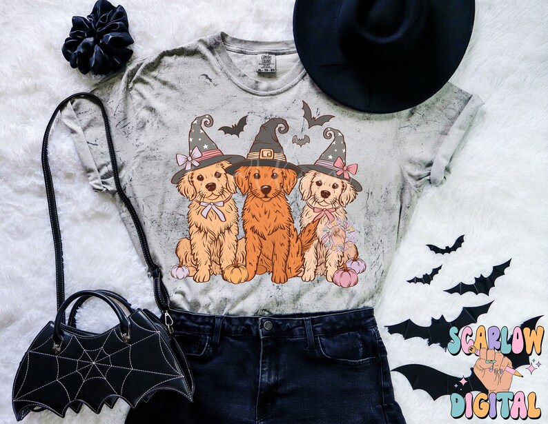 K&ouml;nnte beinhalten: Ein graues T-Shirt mit einem Used-Look zeigt drei Golden Retriever, die Hexenh&uuml;te tragen und K&uuml;rbisse halten. Das Shirt liegt auf einer wei&szlig;en Oberfl&auml;che mit einem schwarzen Hut und einer schwarzen Handtasche mit Spinnennetz-Design.