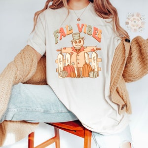 Op de afbeelding: Een wit T-shirt met de tekst "Fall Vibes" in kleurrijke letters. Een cartoon-vogelverschrikker met een hoed en een pompoen in elke hand staat voor een wit hek met pompoenen aan beide kanten.