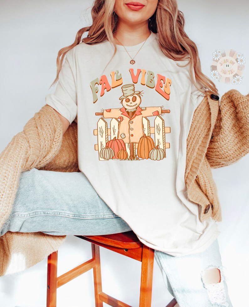 Op de afbeelding: Een wit T-shirt met de tekst "Fall Vibes" in kleurrijke letters. Een cartoon-vogelverschrikker met een hoed en een pompoen in elke hand staat voor een wit hek met pompoenen aan beide kanten.