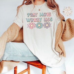 May include: A white t-shirt with the text "Pray More Worry Less" in a retro font with pink, blue, and purple flowers underneath.
