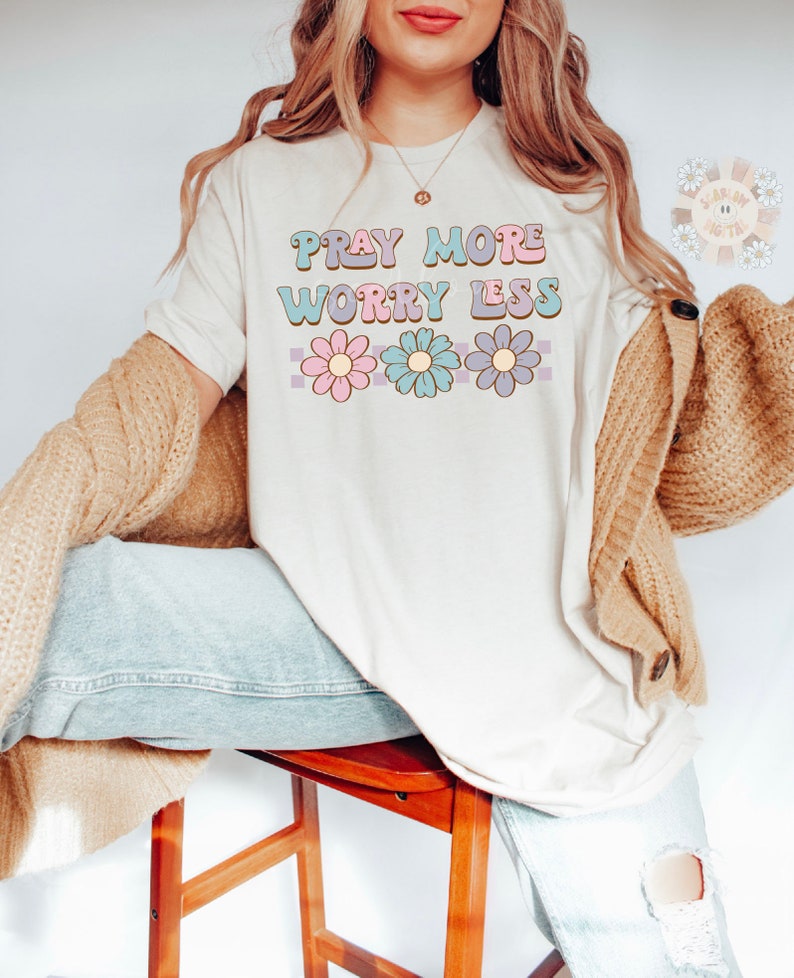 May include: A white t-shirt with the text "Pray More Worry Less" in a retro font with pink, blue, and purple flowers underneath.