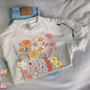 May include: A white t-shirt with a floral print featuring a variety of flowers in shades of pink, yellow, orange, and purple. The text "SCARLOWDIGITAL.COM" is printed on the bottom right corner of the shirt.