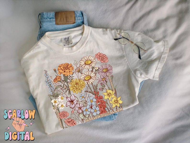 May include: A white t-shirt with a floral print featuring a variety of flowers in shades of pink, yellow, orange, and purple. The text "SCARLOWDIGITAL.COM" is printed on the bottom right corner of the shirt.