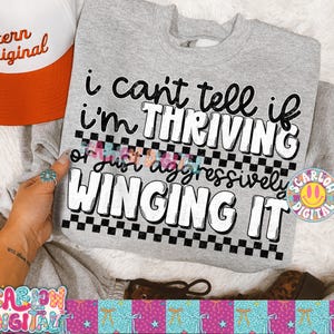 May include: A heather gray sweatshirt with the text "i can't tell if I'm THRIVING or just aggressively WINGING IT" in black and white. A red and white trucker hat is visible in the upper left corner.