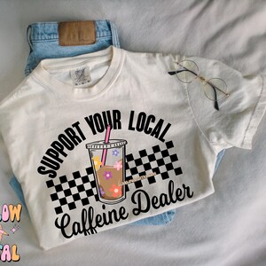 May include: White t-shirt with black text that reads "Support Your Local Caffeine Dealer". The text is in a circle with a checkered pattern and a cartoon iced coffee cup with flowers.