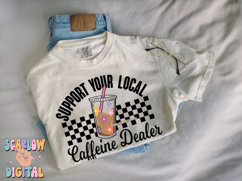 May include: White t-shirt with black text that reads "Support Your Local Caffeine Dealer". The text is in a circle with a checkered pattern and a cartoon iced coffee cup with flowers.