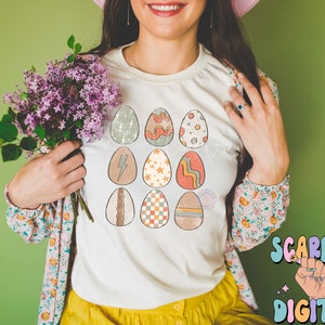 May include: A white t-shirt with a design of 12 colorful Easter eggs. The eggs have different patterns, including stripes, polka dots, and geometric shapes.