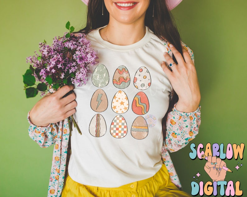 May include: A white t-shirt with a design of 12 colorful Easter eggs. The eggs have different patterns, including stripes, polka dots, and geometric shapes.