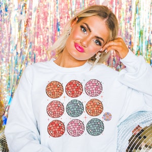 May include: White sweatshirt with a colorful design of nine disco balls. The disco balls are arranged in a 3x3 grid and have a retro, vintage aesthetic.