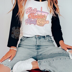 May include: A white and black raglan style t-shirt with the text "Half Human Half Coffee" in a retro font. The shirt features a graphic of a coffee drink. The model is wearing light wash ripped jeans and white sneakers.