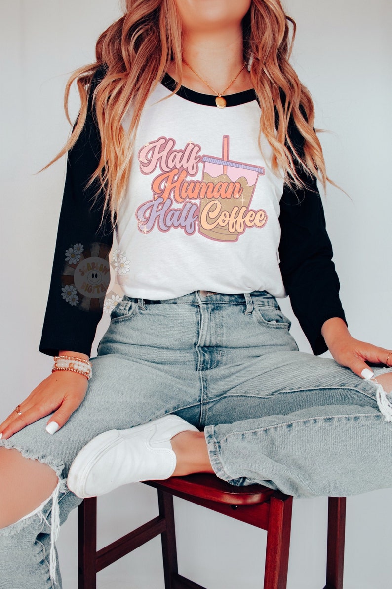 May include: A white and black raglan style t-shirt with the text "Half Human Half Coffee" in a retro font. The shirt features a graphic of a coffee drink. The model is wearing light wash ripped jeans and white sneakers.
