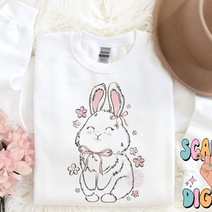 May include: White sweatshirt with a cute cartoon bunny wearing a pink bow and surrounded by pink flowers. The bunny is smiling and has a pink nose.