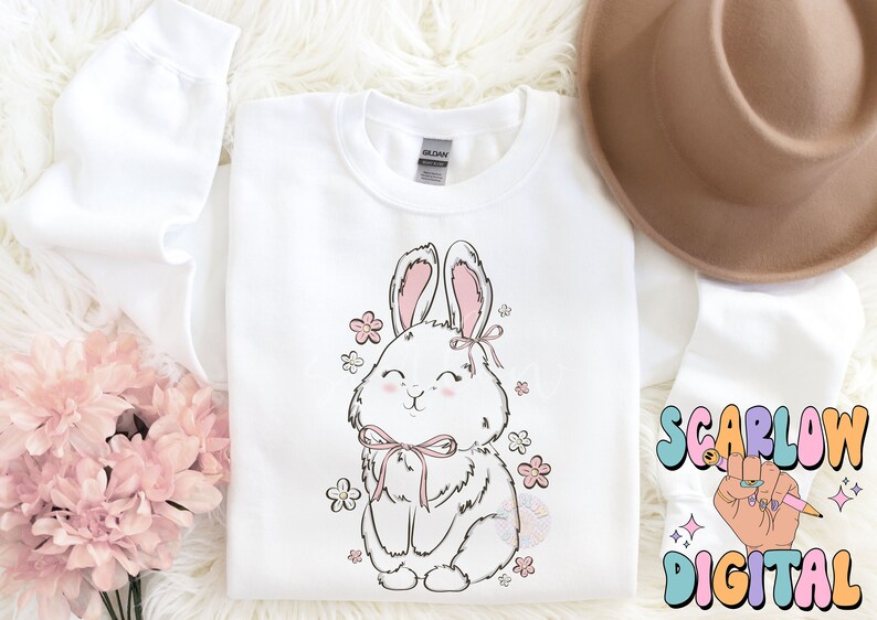 May include: White sweatshirt with a cute cartoon bunny wearing a pink bow and surrounded by pink flowers. The bunny is smiling and has a pink nose.