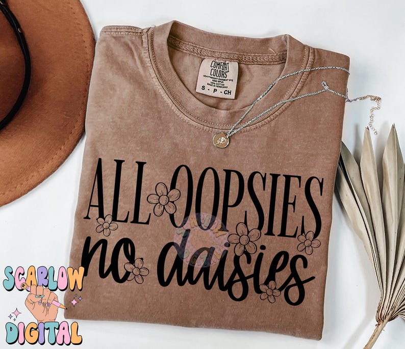Puede incluir: Una camiseta marr&oacute;n con el texto "All oopsies no daisies" en letras negras con peque&ntilde;as ilustraciones de margaritas.