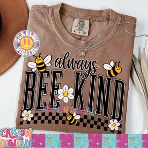 May include: Brown Comfort Colors t-shirt with the phrase "always BEE KIND" in black and white. The design includes bee and daisy graphics. A silver necklace rests on the shirt. The shirt is size S-P-CH.