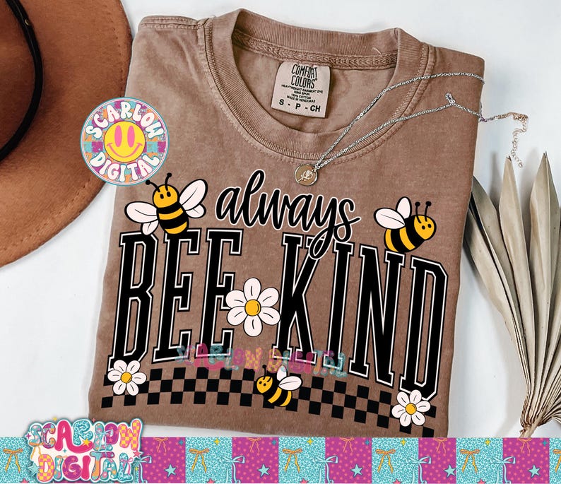May include: Brown Comfort Colors t-shirt with the phrase "always BEE KIND" in black and white. The design includes bee and daisy graphics. A silver necklace rests on the shirt. The shirt is size S-P-CH.