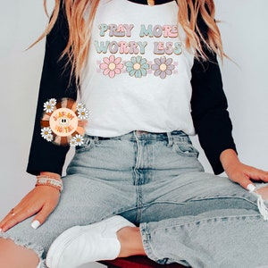 May include: A white and black baseball tee shirt with the text "Pray More Worry Less" in colorful letters and a row of flowers below the text. The model is wearing blue jeans and white sneakers.
