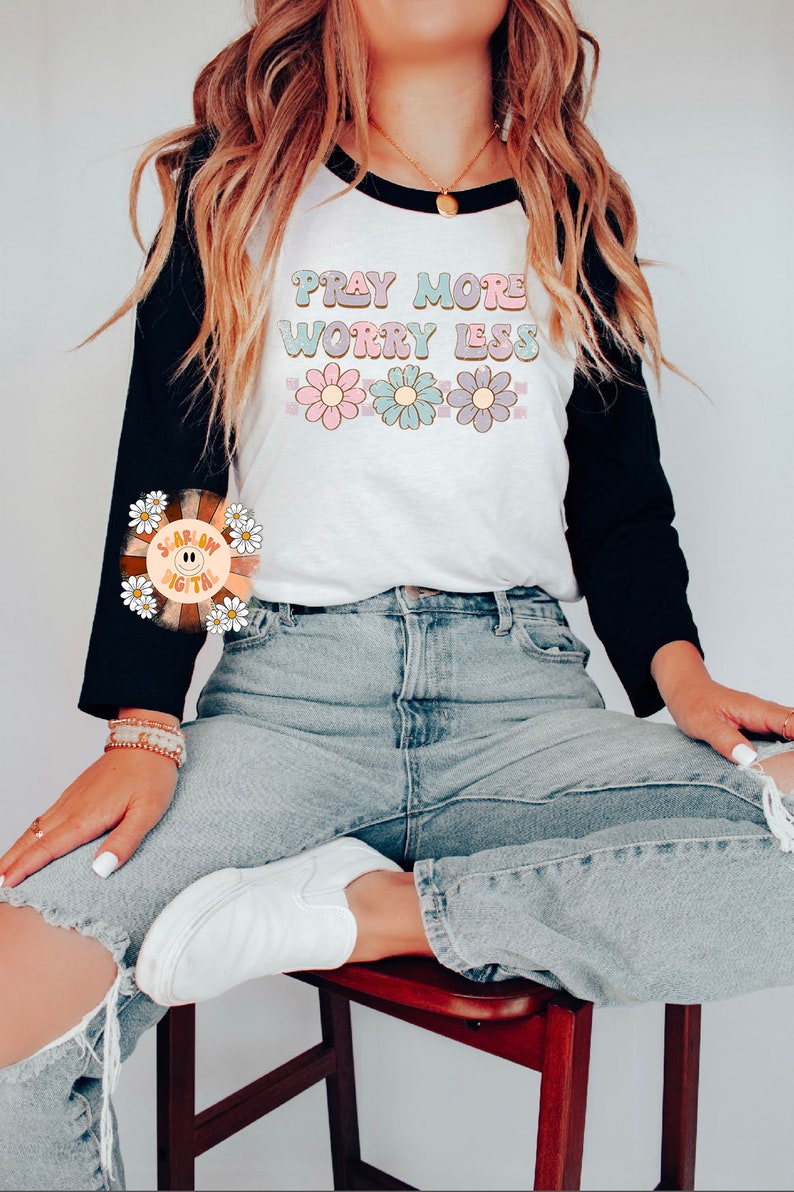 May include: A white and black baseball tee shirt with the text "Pray More Worry Less" in colorful letters and a row of flowers below the text. The model is wearing blue jeans and white sneakers.