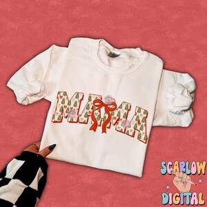 May include: White t-shirt with the word "MAMA" printed in red outlined letters. The letters are filled with a pattern of green Christmas trees and red bows.