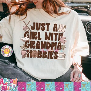 K&ouml;nnte beinhalten: Cremefarbenes T-Shirt mit dem Text "JUST A GIRL WITH GRANDMA HOBBIES" in Braun. Das Shirt zeigt Illustrationen von Stricknadeln, einem Wollkn&auml;uel, einem Buch, einem Notizblock, einem Puzzleteil, einer Topfpflanze, einem Wollkn&auml;uel und einer N&auml;hmaschine.