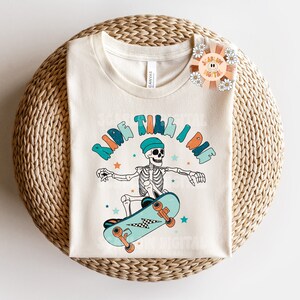 K&ouml;nnte beinhalten: Ein wei&szlig;es T-Shirt mit einem Grafikdruck eines Skeletts, das Skateboard f&auml;hrt, und dem Text "Ride Till I Die".