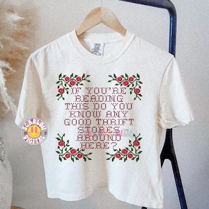 May include: Cream-colored t-shirt with the text "IF YOU'RE READING THIS DO YOU KNOW ANY GOOD THRIFT STORES AROUND HERE?" in a cross-stitch style. The text is framed by red roses and green leaves.