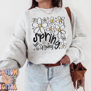 Puede incluir: Sudadera gris claro con la frase "spring has sprung" en letras negras y un dise&ntilde;o floral. Las flores son blancas con centros amarillos. La sudadera tiene mangas largas y cuello redondo.