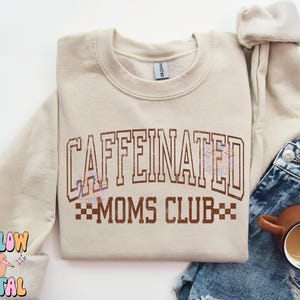 Puede incluir: Una sudadera beige con el texto "CAFFEINATED MOMS CLUB" en letras marrones con un diseño a cuadros debajo. La sudadera tiene cuello redondo y mangas largas.