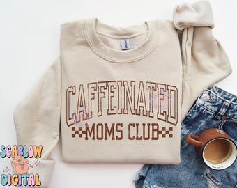 Op de afbeelding: Een beige sweatshirt met de tekst "CAFFEINATED MOMS CLUB" in bruine letters met een geruit ontwerp eronder. Het sweatshirt heeft een ronde hals en lange mouwen.