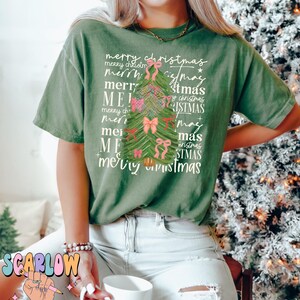 May include: A green t-shirt with a Christmas tree design made of the words "merry christmas" repeated in different sizes and styles. The tree is decorated with pink bows.